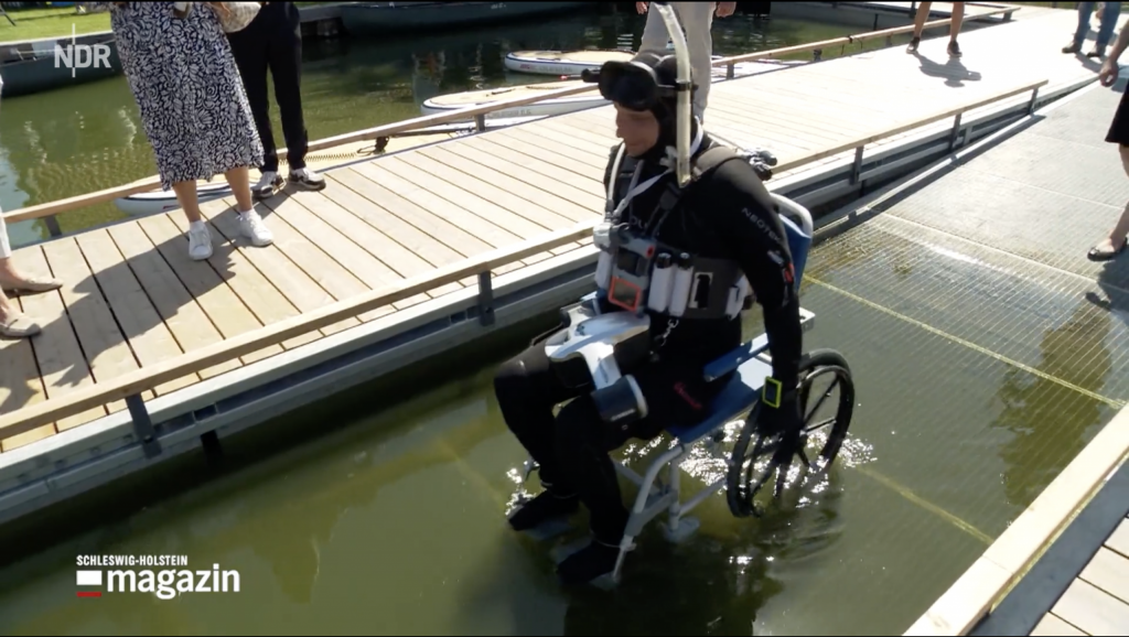 Rollstuhlfahrer in Tauchausrüstung fährt über Rampe ins Wasser